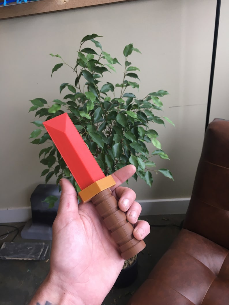May include: A red and brown plastic toy sword with a textured handle. The sword is held in a person's hand.