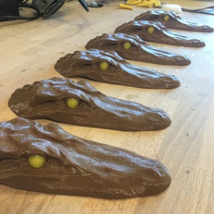 May include: A row of six brown alligator head sculptures with yellow eyes. The sculptures are arranged in a line on a wooden surface.