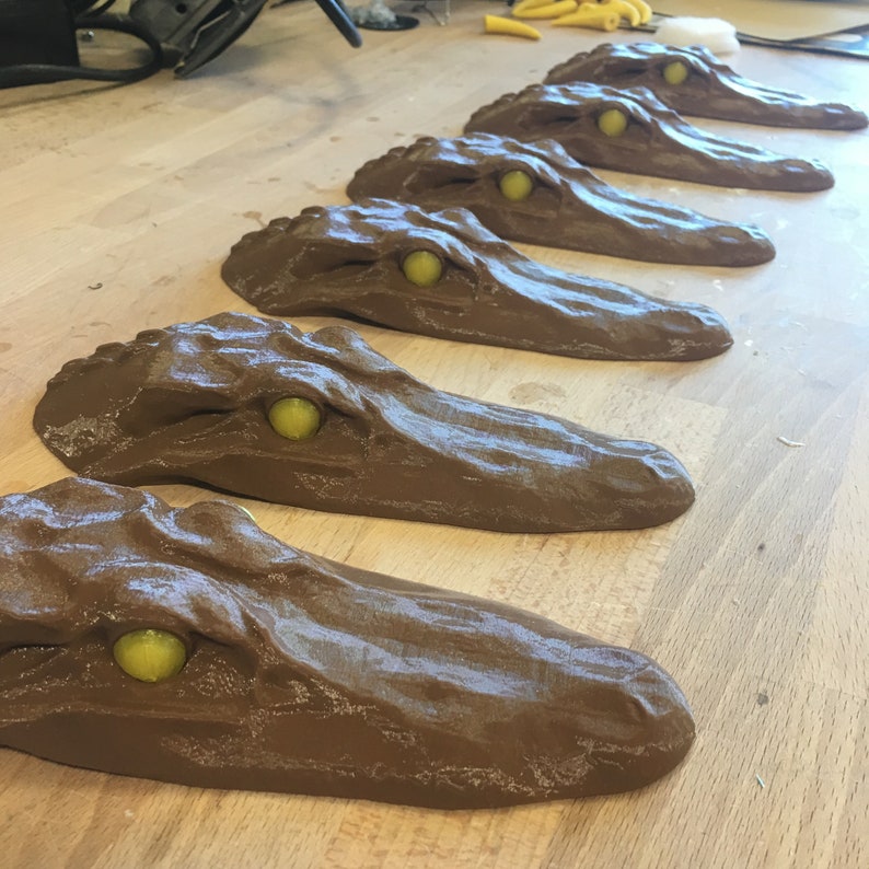 May include: A row of six brown alligator head sculptures with yellow eyes. The sculptures are arranged in a line on a wooden surface.