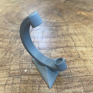 May include: A gray 3D printed stand with a curved arm and a circular opening at the top. The stand is shaped like a triangle and has a textured surface.