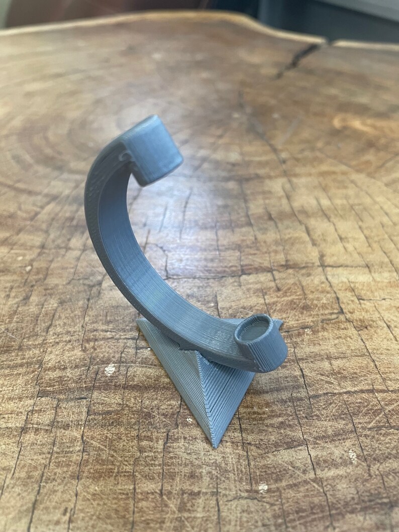 May include: A gray 3D printed stand with a curved arm and a circular opening at the top. The stand is shaped like a triangle and has a textured surface.