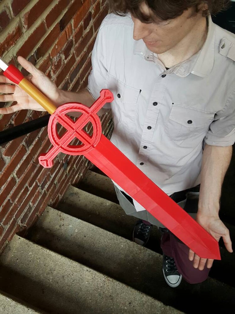 Op de afbeelding: Een rood zwaard met een ronde bescherming met een zwart kruisontwerp. Het zwaard heeft een wit en gouden handvat.