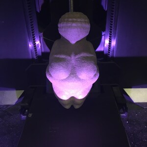 Puede incluir: Una figurita de mujer impresa en 3D, impresa en color gris claro, con un brillo p&uacute;rpura que emana del objeto.