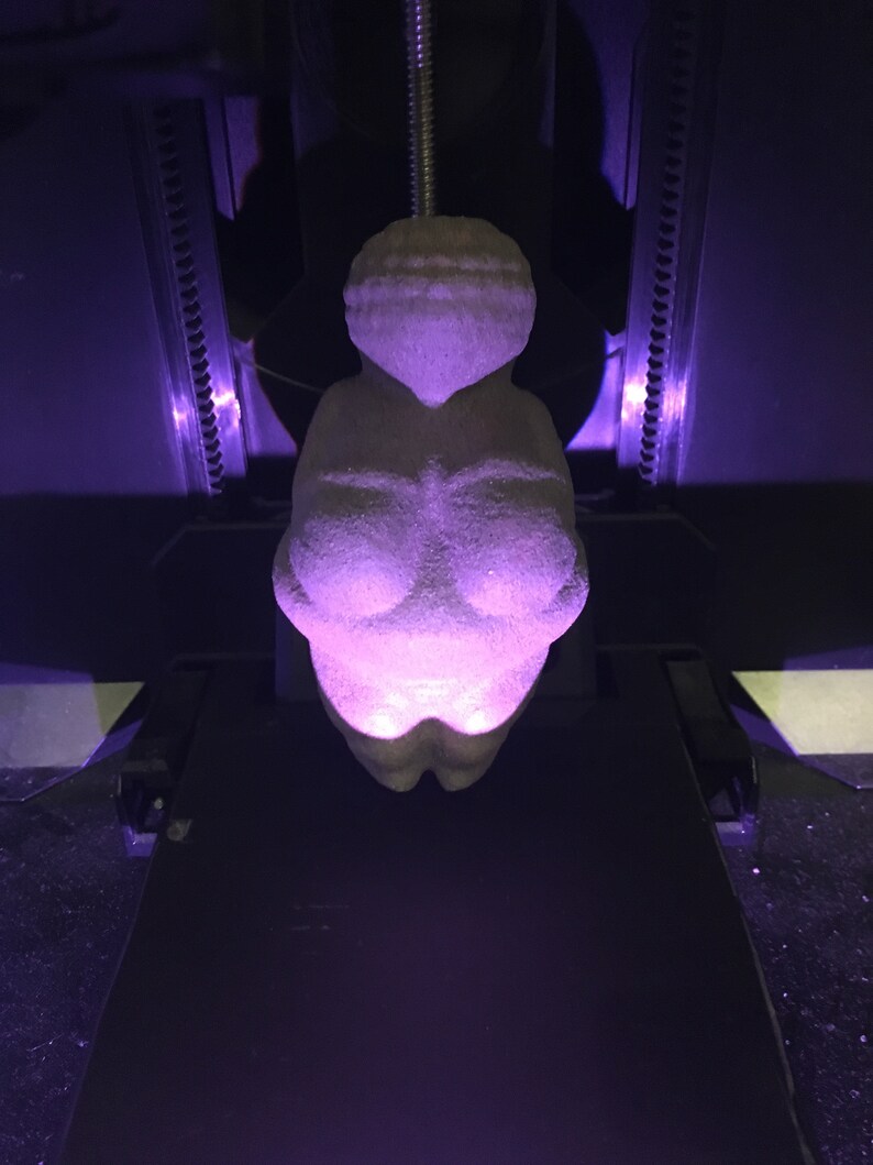 Puede incluir: Una figurita de mujer impresa en 3D, impresa en color gris claro, con un brillo p&uacute;rpura que emana del objeto.