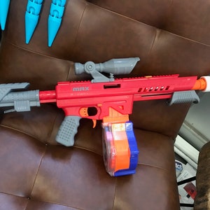 May include: A red and gray toy gun with a black stock and a clear orange and blue magazine. The gun has a scope on top and the word "MAX" printed on the side.