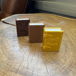 May include: Three small decorative boxes with a wood grain texture. The boxes are brown, brown, and gold. The gold box has the words "Spell Book" printed on the front.