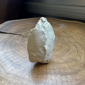 May include: A white, irregularly shaped stone with a rough, textured surface. The stone is sitting on a wooden surface.