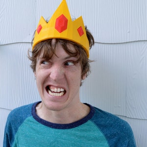 May include: A person wearing a yellow crown with red gems on top. The crown is made of plastic and has a pointed top. The person is looking to the right and has a serious expression on their face.