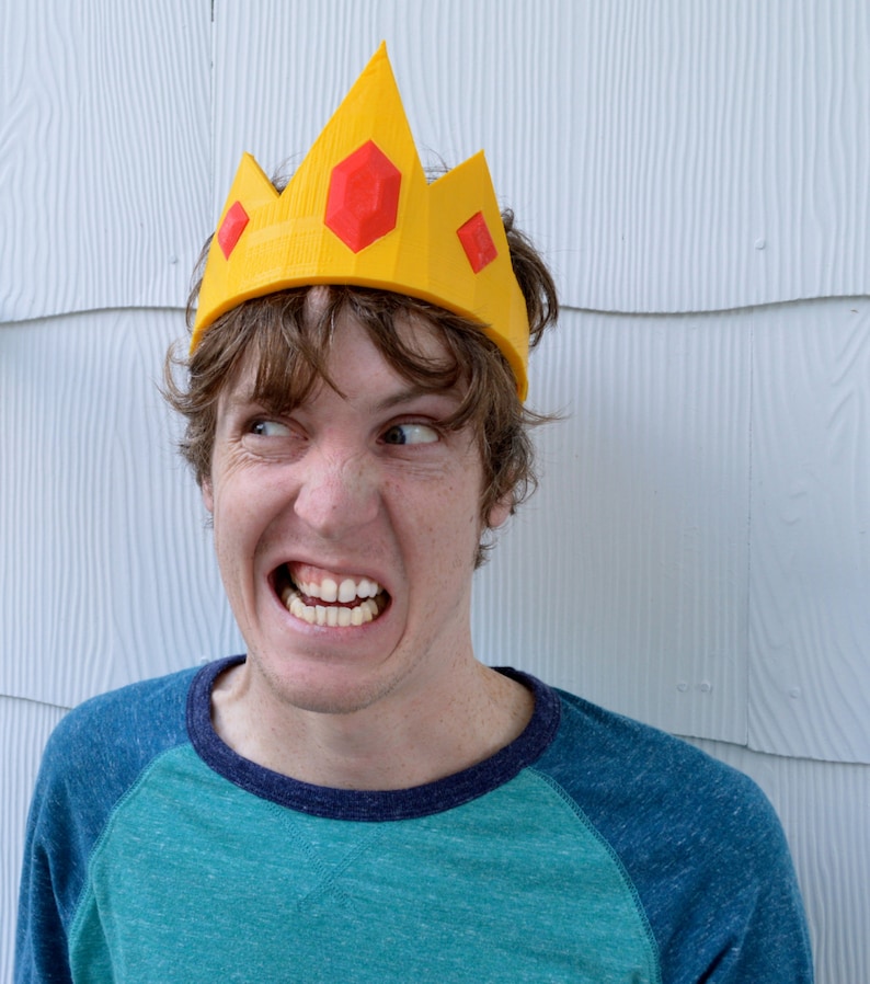 May include: A person wearing a yellow crown with red gems on top. The crown is made of plastic and has a pointed top. The person is looking to the right and has a serious expression on their face.