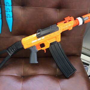 May include: A black and orange toy gun with a black magazine and a black stock. The gun has a black barrel with a orange tip and a black handle. The gun is sitting on a brown leather surface.