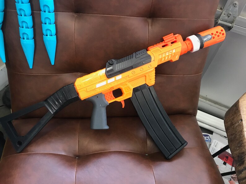 May include: A black and orange toy gun with a black magazine and a black stock. The gun has a black barrel with a orange tip and a black handle. The gun is sitting on a brown leather surface.