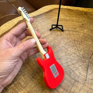 May include: A red electric guitar with a wooden neck and silver tuning pegs. The guitar is being held in a hand and is on a wooden surface.