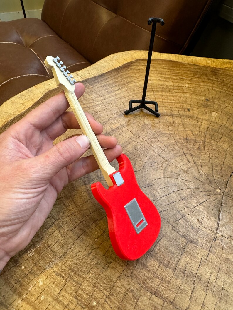 May include: A red electric guitar with a wooden neck and silver tuning pegs. The guitar is being held in a hand and is on a wooden surface.