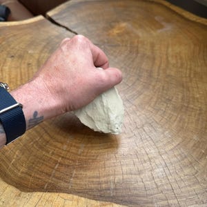 May include: A hand holding a light brown, irregularly shaped rock on a wooden surface.