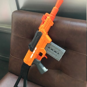 May include: An orange and gray toy gun with a black handle and stock. The gun has a magazine and a barrel with a series of holes. The text "AGON-PRO" is visible on the side of the gun.