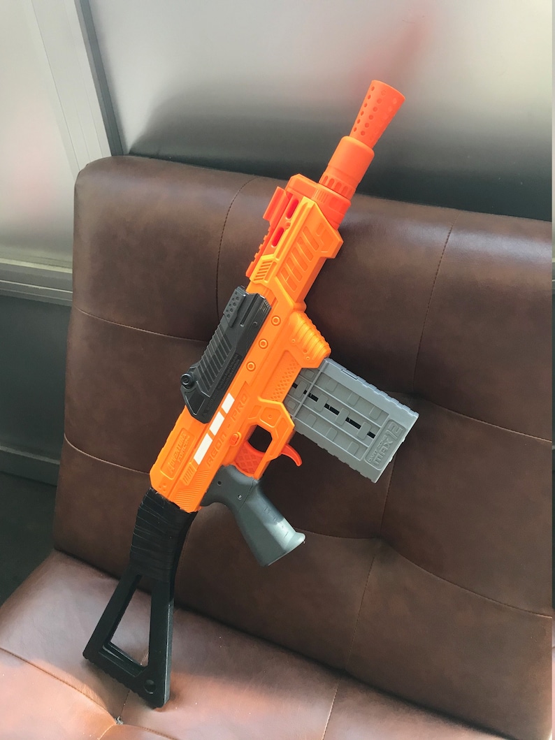 May include: An orange and gray toy gun with a black handle and stock. The gun has a magazine and a barrel with a series of holes. The text "AGON-PRO" is visible on the side of the gun.
