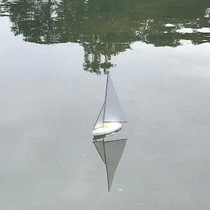 Puede incluir: Un velero blanco con un m&aacute;stil y una vela negros flota en una masa de agua tranquila. El velero se refleja en el agua.