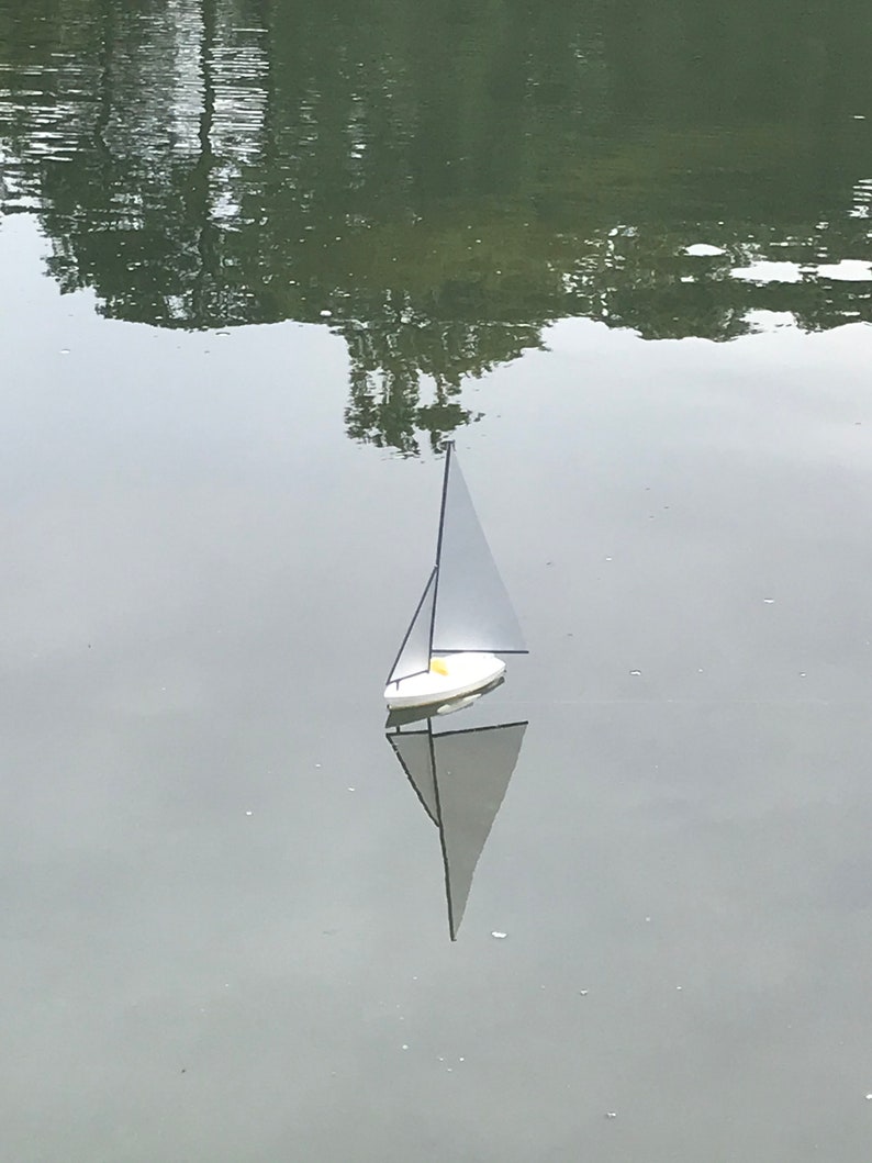 Puede incluir: Un velero blanco con un m&aacute;stil y una vela negros flota en una masa de agua tranquila. El velero se refleja en el agua.
