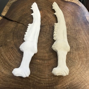 May include: Two white, ceramic, stylized, shark jaw-shaped daggers on a wooden surface.