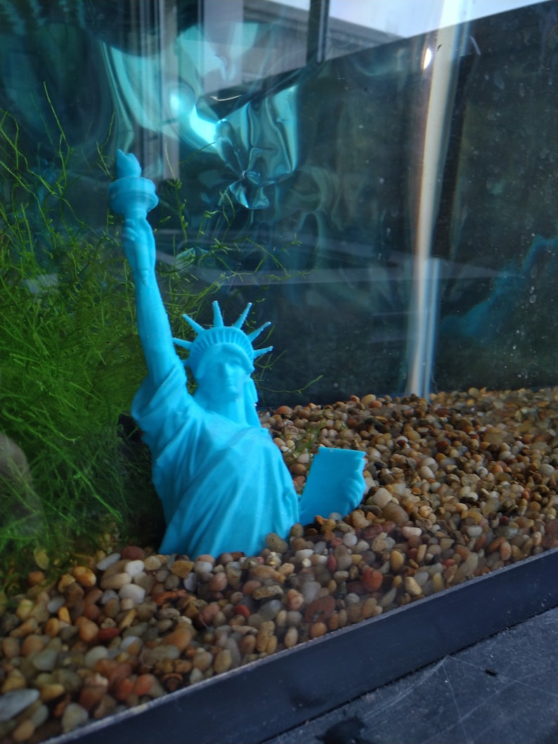 K&ouml;nnte beinhalten: Eine blaue Plastikstatue der Freiheitsstatue ist teilweise in einem Aquarium mit kleinen, glatten, braunen, wei&szlig;en und grauen Steinen versunken. Die Statue h&auml;lt eine Fackel in der rechten Hand und ein Buch in der linken Hand.