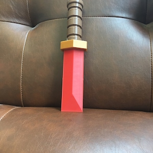 May include: A red plastic toy sword with a brown plastic handle. The sword has a gold-colored hilt.
