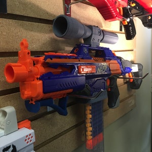 Może przedstawiać: Niebiesko-pomarańczowy blaster Nerf z przezroczystym magazynkiem i przymocowanym czarnym celownikiem. Blaster jest zamontowany na ścianie.