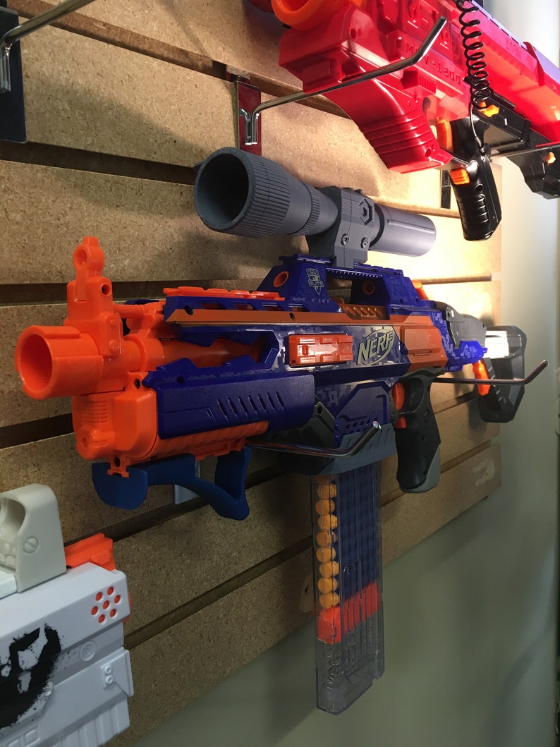Może przedstawiać: Niebiesko-pomarańczowy blaster Nerf z przezroczystym magazynkiem i przymocowanym czarnym celownikiem. Blaster jest zamontowany na ścianie.