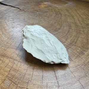 May include: A white ceramic sculpture of a rock, with a smooth, rounded surface. The sculpture is sitting on a wooden surface.