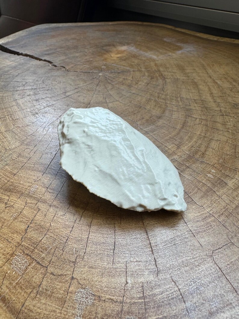 May include: A white ceramic sculpture of a rock, with a smooth, rounded surface. The sculpture is sitting on a wooden surface.