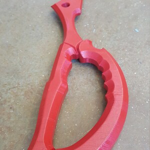 May include: A red plastic prop sword shaped like a pair of scissors. The sword has a sharp point and a curved handle.