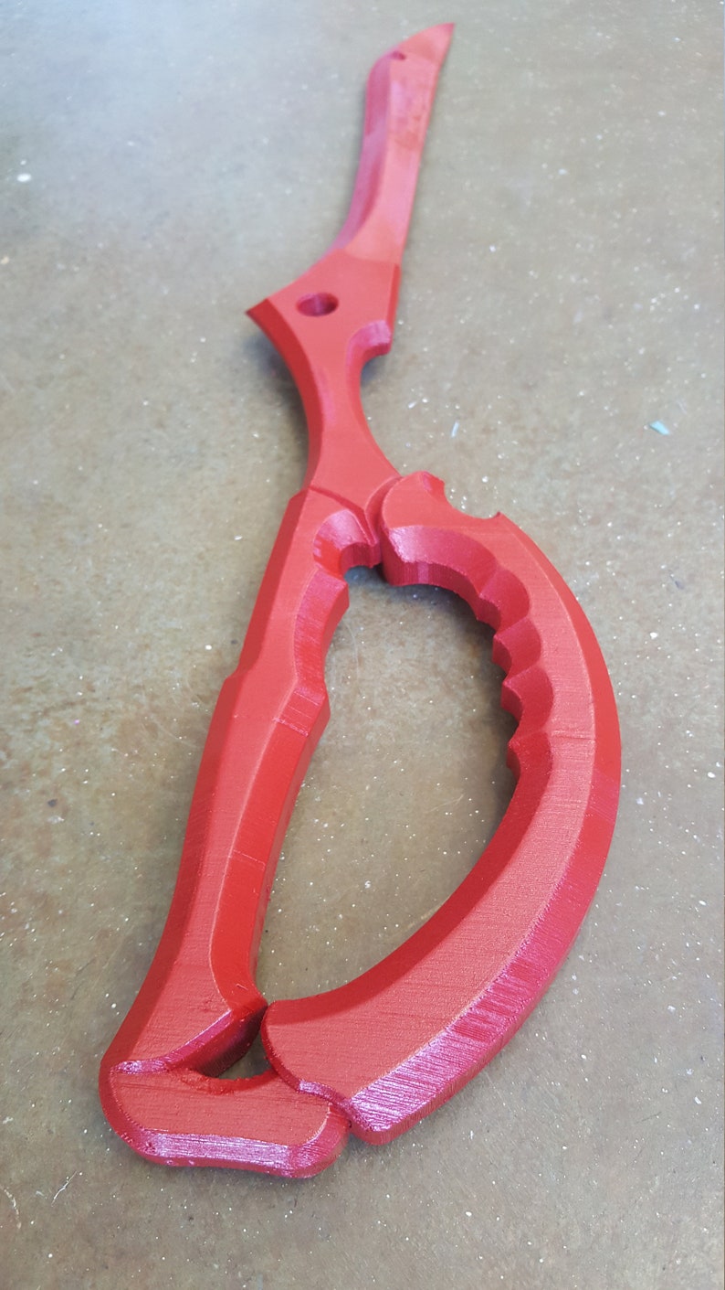 May include: A red plastic prop sword shaped like a pair of scissors. The sword has a sharp point and a curved handle.