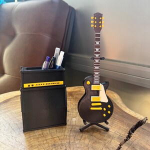 May include: A black and yellow electric guitar model on a stand next to a black amplifier with a pen holder. The amplifier has a yellow stripe and a row of knobs.