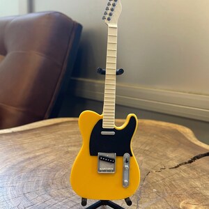 May include: A yellow electric guitar with a black pickguard and a white neck. The guitar is on a black stand.