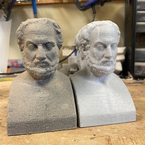 May include: Two 3D printed busts of a man with a beard, one is gray and the other is white. The busts are on a wooden surface.
