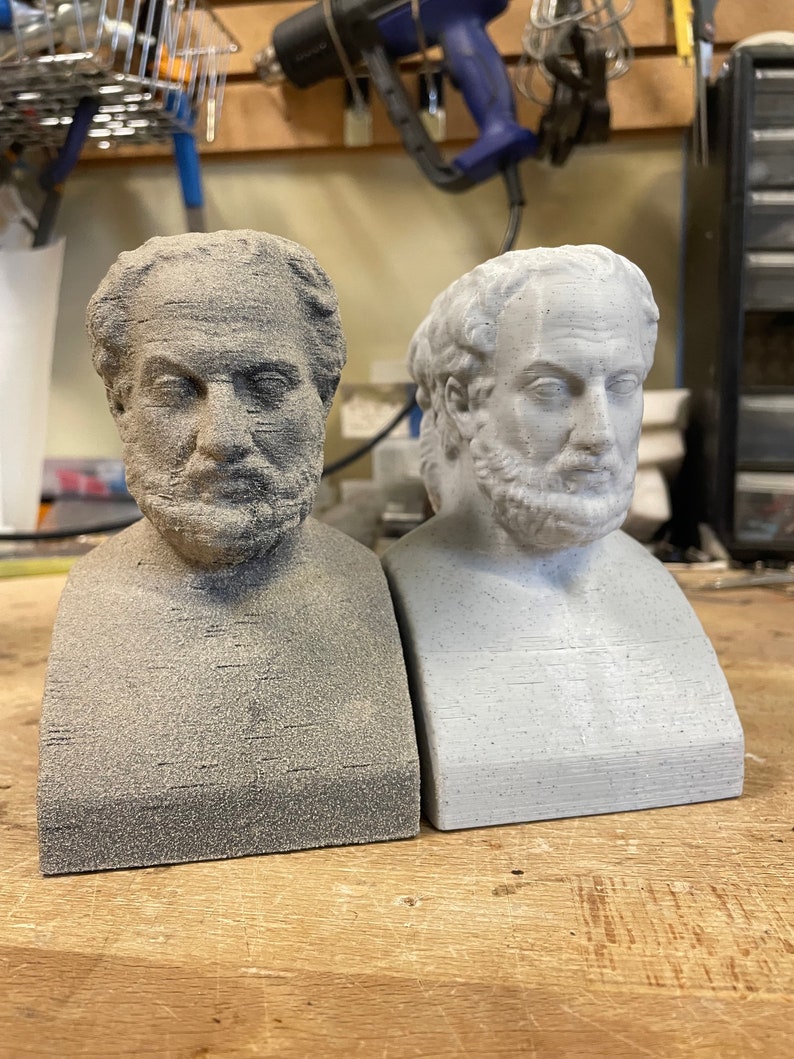 May include: Two 3D printed busts of a man with a beard, one is gray and the other is white. The busts are on a wooden surface.
