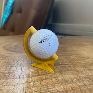 May include: A gold-colored golf ball holder with a golf ball inside. The golf ball is white with black text that reads "TiTech Titanium 2".
