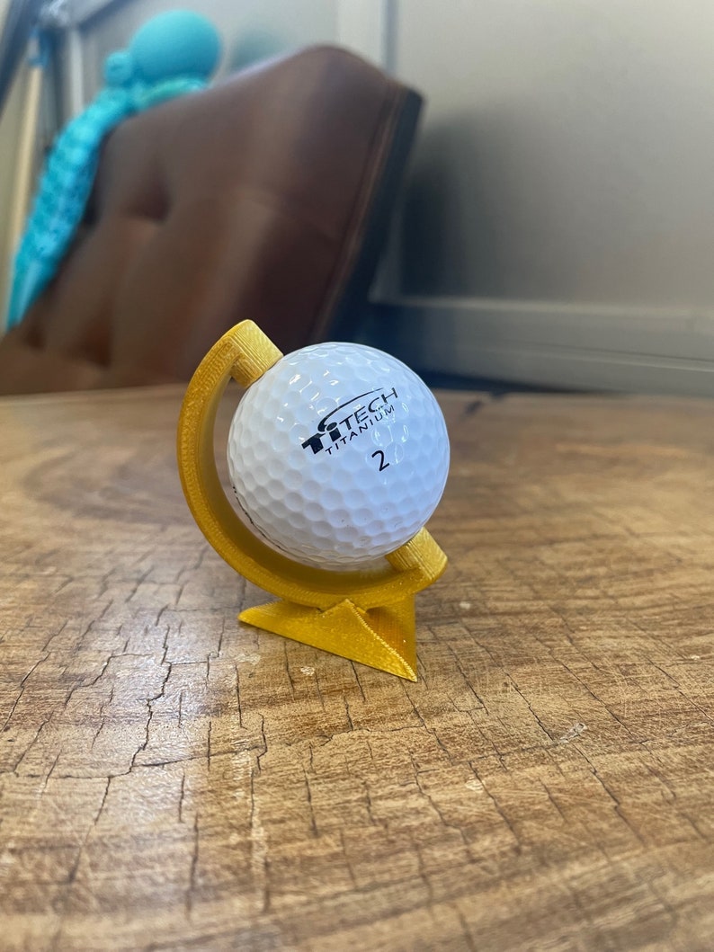 May include: A gold-colored golf ball holder with a golf ball inside. The golf ball is white with black text that reads "TiTech Titanium 2".