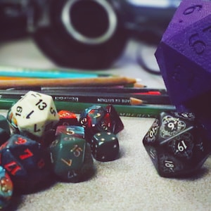 May include: A collection of colorful dice, including a purple, multi-sided die with numbers on each side, and a black die with white numbers and a decorative design. The dice are on a table with pencils and other supplies.