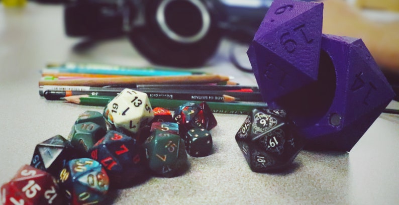 May include: A collection of colorful dice, including a purple, multi-sided die with numbers on each side, and a black die with white numbers and a decorative design. The dice are on a table with pencils and other supplies.