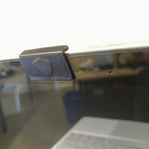 May include: A black plastic webcam cover with a shield design, attached to a laptop screen.