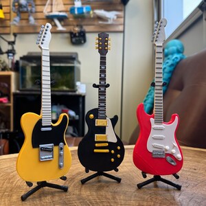 May include: Three miniature electric guitars on stands. The guitars are yellow, black, and red. The yellow guitar has a black pickguard. The black guitar has a white pickguard. The red guitar has a white pickguard.