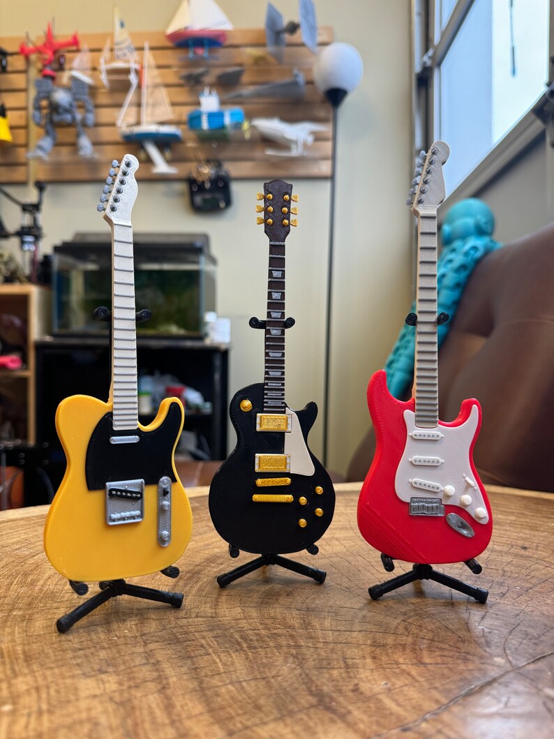 May include: Three miniature electric guitars on stands. The guitars are yellow, black, and red. The yellow guitar has a black pickguard. The black guitar has a white pickguard. The red guitar has a white pickguard.