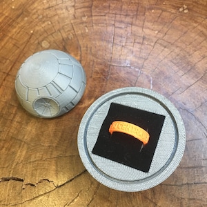 May include: A 3D printed ring box shaped like the Death Star from Star Wars. The box is open and a bright orange ring with the word "GENERAL" inscribed on it is inside.