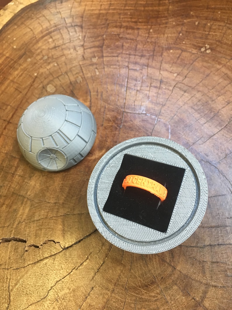 May include: A 3D printed ring box shaped like the Death Star from Star Wars. The box is open and a bright orange ring with the word "GENERAL" inscribed on it is inside.
