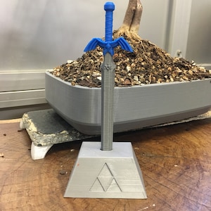May include: A blue plastic sword with a winged handle and a triangular base with a symbol. The sword is on a wooden surface.