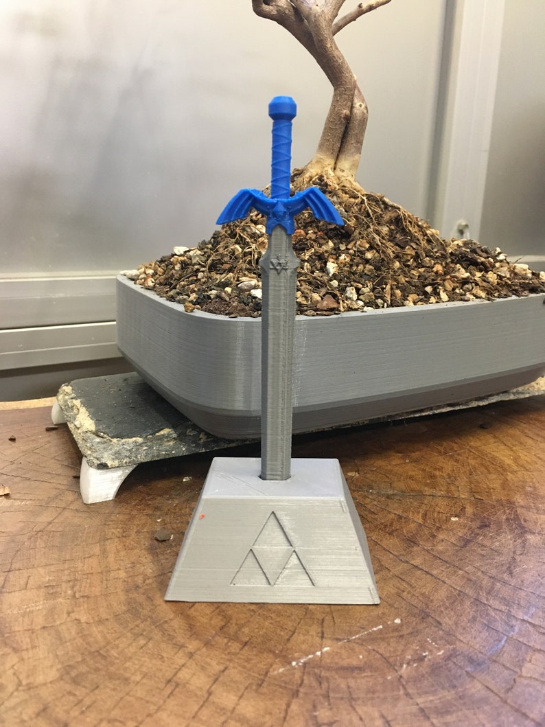 May include: A blue plastic sword with a winged handle and a triangular base with a symbol. The sword is on a wooden surface.
