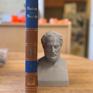 May include: A brown and blue hardcover book with gold lettering titled "Herodotus - Thucydides" and the number "6" on the spine. The book is part of the "Great Books" series. A gray bust of a man with a beard is in the background.