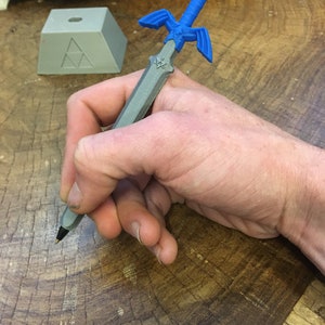May include: A gray 3D printed pen shaped like the Master Sword from the Legend of Zelda video game series. The pen has a blue handle and a silver blade.