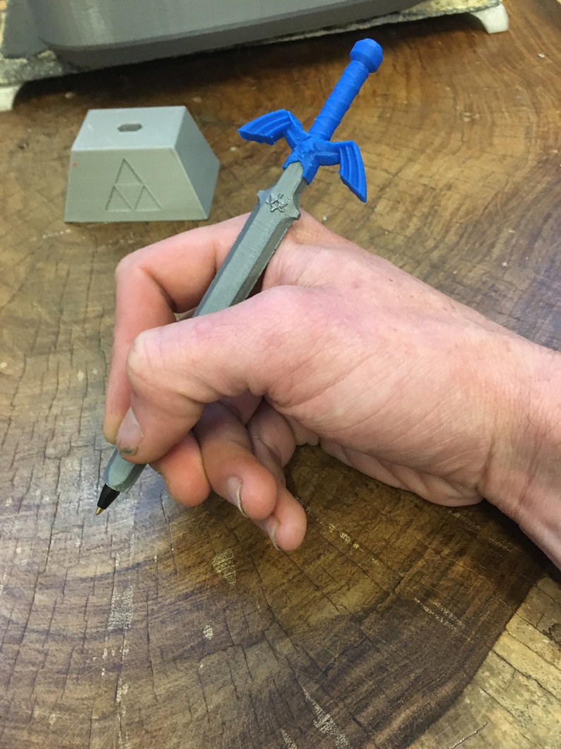 May include: A gray 3D printed pen shaped like the Master Sword from the Legend of Zelda video game series. The pen has a blue handle and a silver blade.