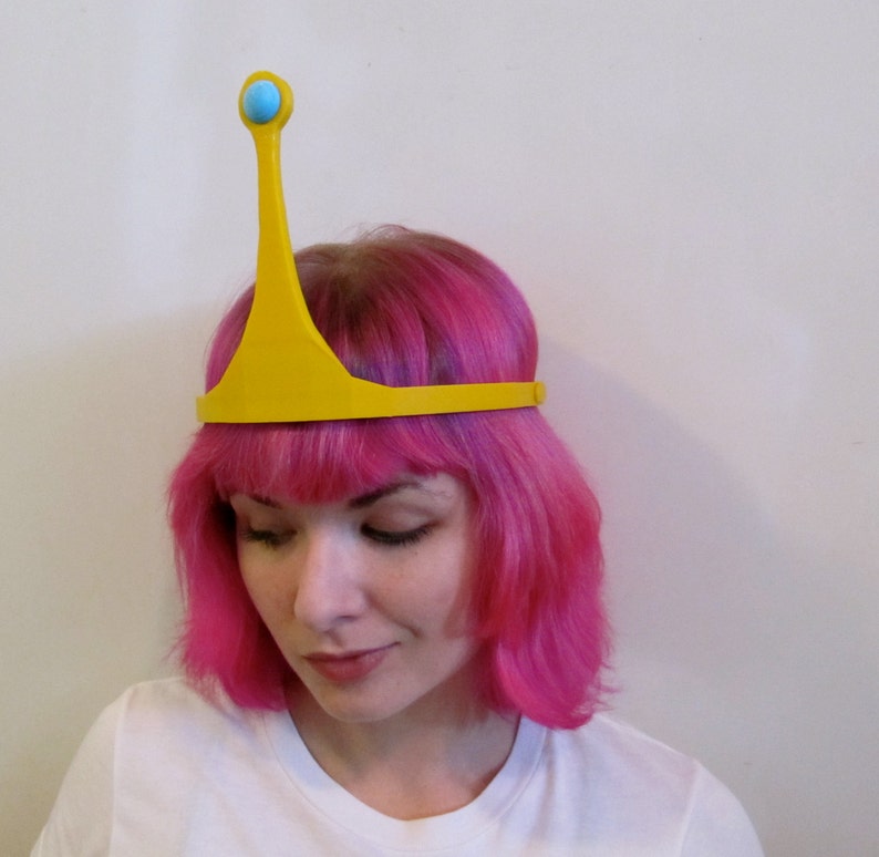 May include: A yellow crown with a blue gem on top. The crown is worn by a person with pink hair.